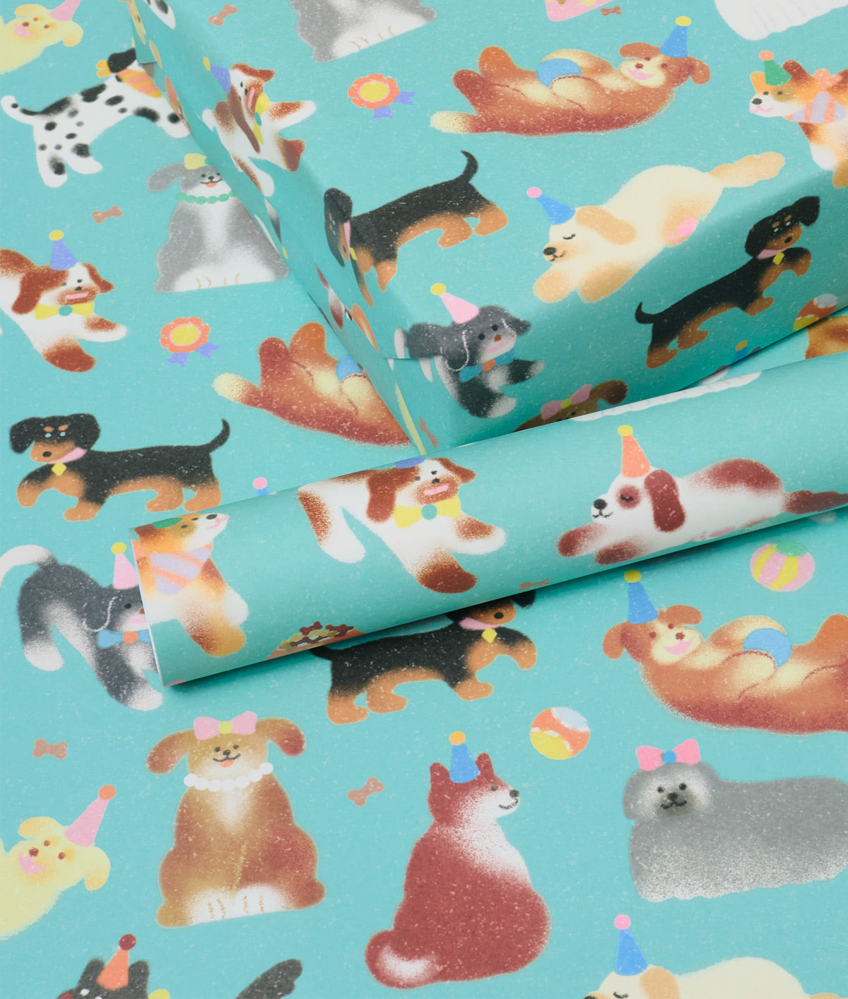 A close-up of turquoise wrapping paper printed with a playful pattern of various dog breeds wearing party hats, shown with one roll partially unrolled.