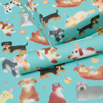 A close-up of turquoise wrapping paper printed with a playful pattern of various dog breeds wearing party hats, shown with one roll partially unrolled.